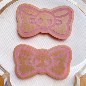May include: Two pink and tan bow-shaped embellishments with a skull design in the center. The bows have a layered effect with a pink outline and a tan interior. The skull is tan with a detailed design. The bows are on a white surface.