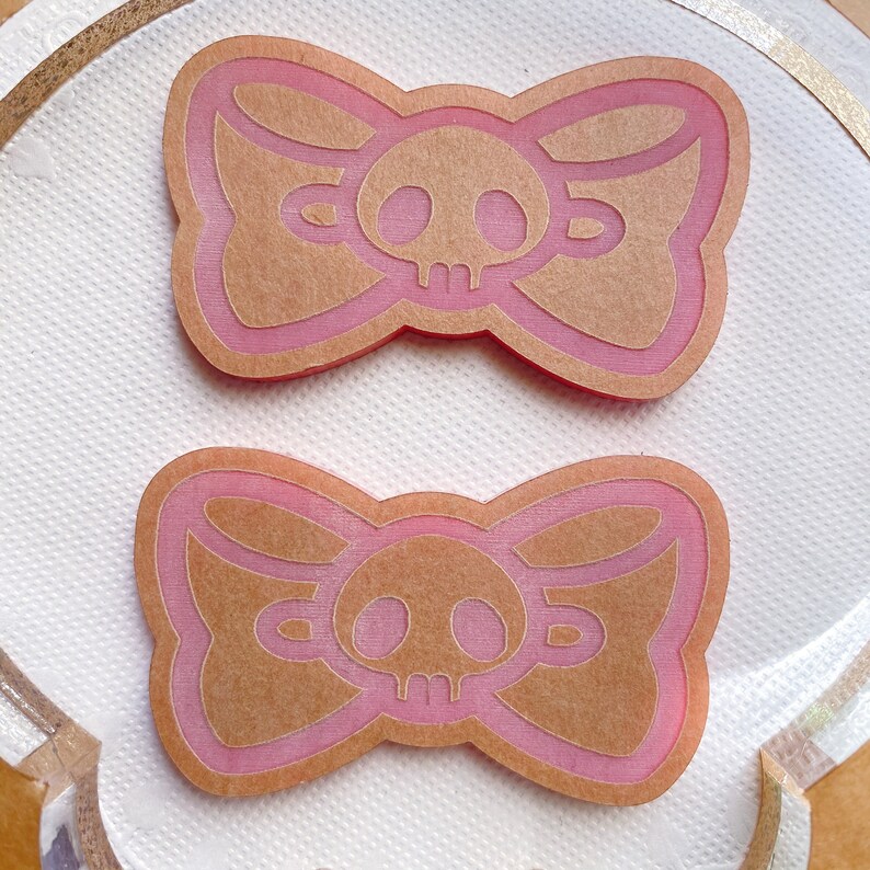 May include: Two pink and tan bow-shaped embellishments with a skull design in the center. The bows have a layered effect with a pink outline and a tan interior. The skull is tan with a detailed design. The bows are on a white surface.