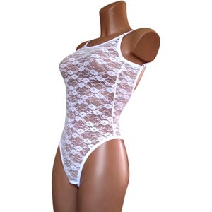 Może przedstawiać: Białe koronkowe body z okrągłym dekoltem i cienkimi ramiączkami. Jednoczęściowy element garderoby ma kwiatowy wzór koronki i wysoki krój nogawek. Body jest prezentowane na manekinie na białym tle.