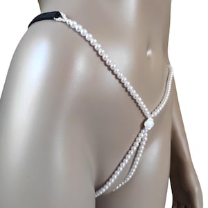 Op de afbeelding: Witte parelstring met een zwarte elastische tailleband. De string heeft een delicaat ontwerp van kleine, ronde parels die aan elkaar zijn geregen en een decoratief patroon vormen. Een klein, wit bloemenaccent bevindt zich aan de voorkant.