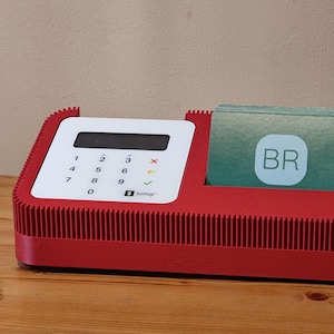 Könnte beinhalten: Ein rotes elektronisches Gerät mit einer weißen Tastatur und einem Schlitz, der einen Stapel Karten enthält. Die Tastatur hat Zahlen und Funktionstasten. Die Karten tragen die Buchstaben "BR". Das Gerät steht auf einer Holzoberfläche.