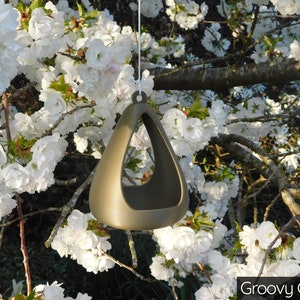 May include: A gold-colored ceramic bird feeder shaped like a triangle with a hole in the center. The feeder is hanging from a tree branch with white blossoms.