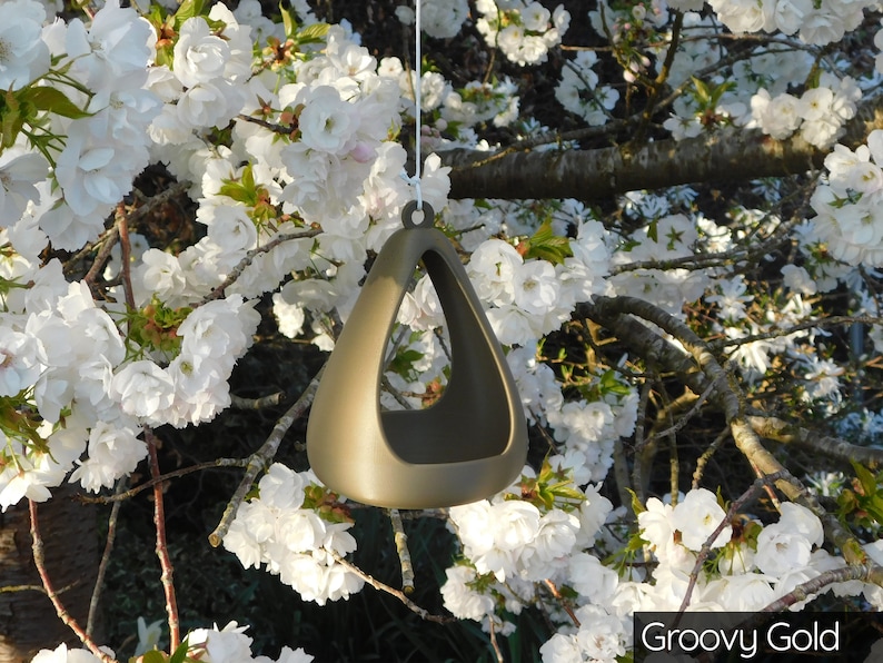 May include: A gold-colored ceramic bird feeder shaped like a triangle with a hole in the center. The feeder is hanging from a tree branch with white blossoms.