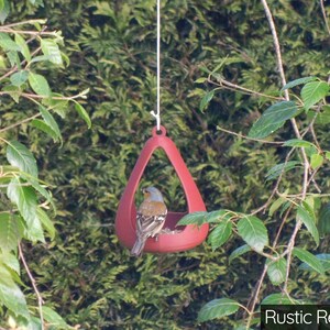 May include: A rustic red bird feeder shaped like a teardrop hangs from a tree branch. A small bird is perched inside the feeder.