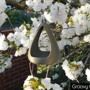 May include: A gold-colored ceramic bird feeder shaped like a triangle with a hole in the center. The feeder is hanging from a tree branch with white blossoms.