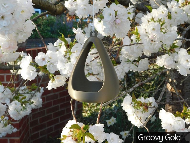 May include: A gold-colored ceramic bird feeder shaped like a triangle with a hole in the center. The feeder is hanging from a tree branch with white blossoms.
