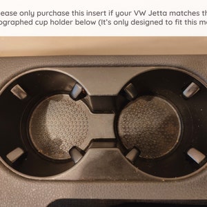 May include: Close-up of a black plastic cup holder with two empty cup slots. The cup holder has a textured surface and is designed to fit a specific car model.