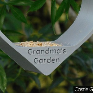 May include: A gray bird feeder shaped like a heart with the words "Grandma's Garden" engraved on the front. The feeder is filled with birdseed and is hanging from a tree branch. The feeder is painted in a color called "Castle Grey".