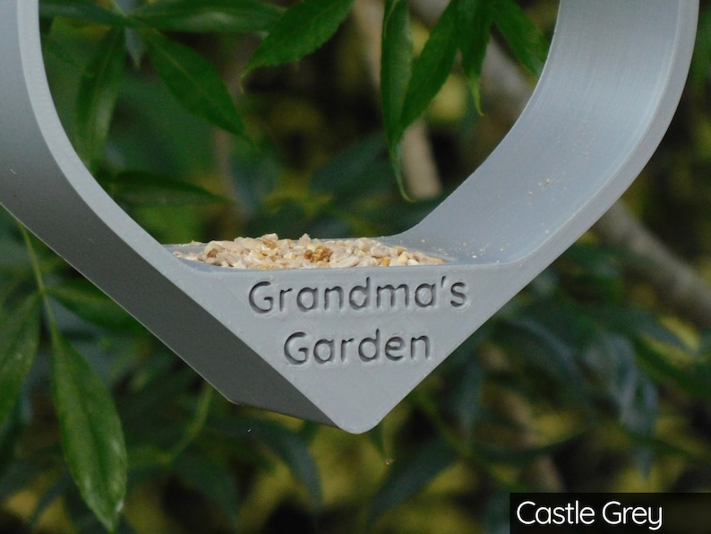 May include: A gray bird feeder shaped like a heart with the words "Grandma's Garden" engraved on the front. The feeder is filled with birdseed and is hanging from a tree branch. The feeder is painted in a color called "Castle Grey".