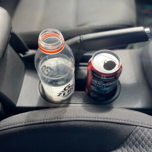 May include: A clear plastic water bottle with a red and white label and a silver can of Rockstar Revolt energy drink in a car's cup holder.