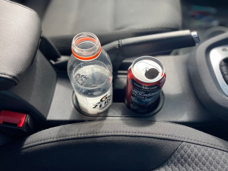 May include: A clear plastic water bottle with a red and white label and a silver can of Rockstar Revolt energy drink in a car's cup holder.