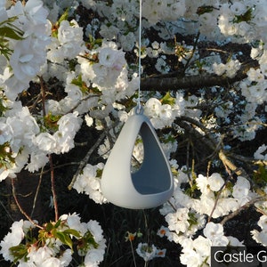 May include: A gray ceramic hanging planter with a triangular shape and a hole in the bottom. The planter is hanging from a white string in front of a tree with white blossoms.