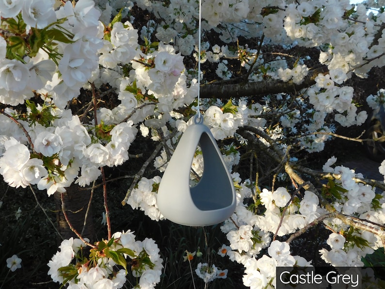 May include: A gray ceramic hanging planter with a triangular shape and a hole in the bottom. The planter is hanging from a white string in front of a tree with white blossoms.