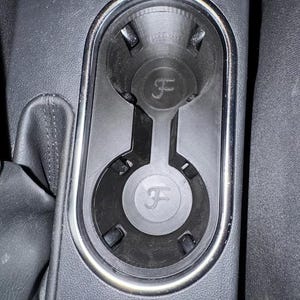 May include: A black car cup holder with a silver trim. The cup holder has two circular compartments and a center divider. Each compartment features a raised "F" logo.