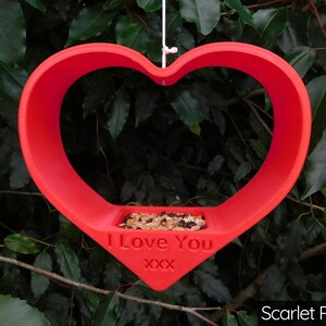 May include: A red heart-shaped bird feeder with the words "I Love You XXX" printed on the front. The feeder is filled with birdseed.