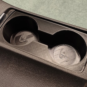 May include: Close-up of a black car cup holder with two circular indentations. Each indentation has a raised "3S" logo.