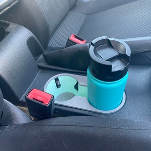 May include: A turquoise travel mug with a black lid sits in a gray cup holder in a car. The cup holder is located in the center console between the driver and passenger seats.