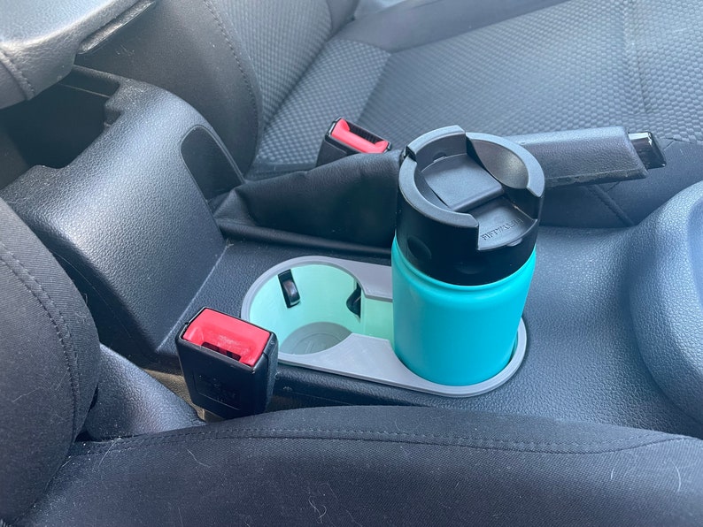 May include: A turquoise travel mug with a black lid sits in a gray cup holder in a car. The cup holder is located in the center console between the driver and passenger seats.