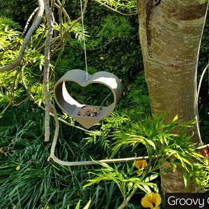 May include: A gray heart-shaped bird feeder hanging from a tree branch. The feeder is filled with birdseed. The text "Groovy Gold" is visible in the lower right corner of the image.