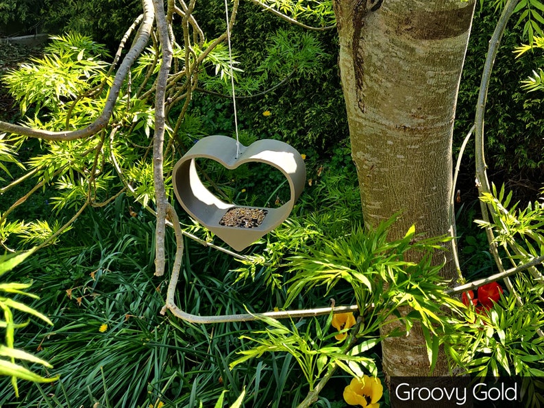 May include: A gray heart-shaped bird feeder hanging from a tree branch. The feeder is filled with birdseed. The text "Groovy Gold" is visible in the lower right corner of the image.