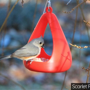 May include: A small bird, a chickadee, is perched on the edge of a bright red, triangular bird feeder. The feeder is hanging from a branch and has a hole in the top for birds to enter.