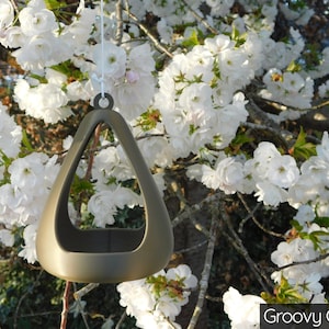May include: A gold-colored, triangular bird feeder hangs from a tree branch. The feeder has a rounded, open top and a smooth, metallic finish. The feeder is hanging in front of a tree with white blossoms.