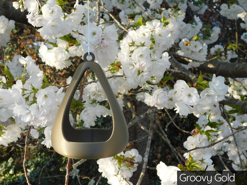 May include: A gold-colored, triangular bird feeder hangs from a tree branch. The feeder has a rounded, open top and a smooth, metallic finish. The feeder is hanging in front of a tree with white blossoms.