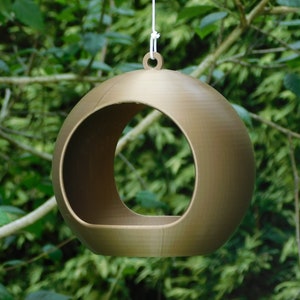 May include: A gold-colored, round bird feeder with a large opening hangs from a white string. The feeder is suspended in front of a green leafy background.