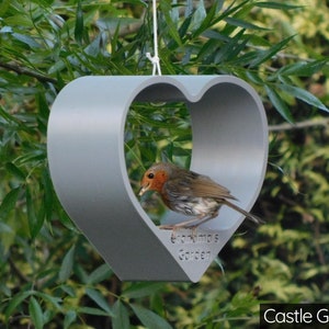 May include: A gray heart-shaped bird feeder with the text "Grandma's Garden" hanging from a tree branch. A robin is perched on the feeder, eating seeds.