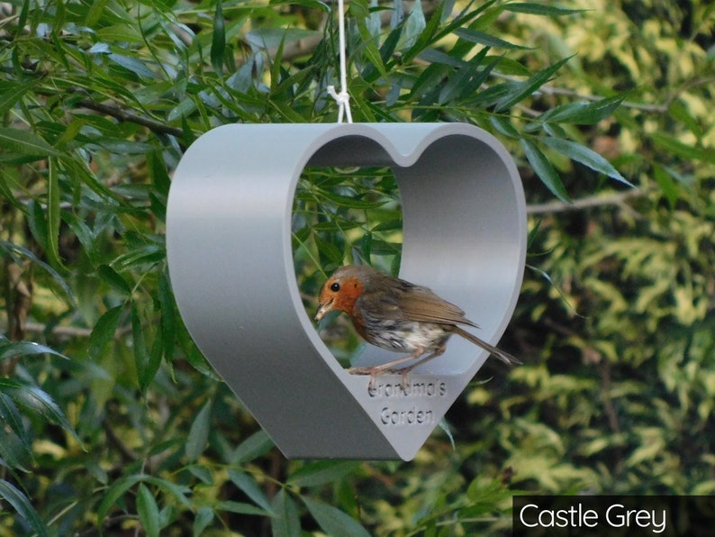 May include: A gray heart-shaped bird feeder with the text "Grandma's Garden" hanging from a tree branch. A robin is perched on the feeder, eating seeds.
