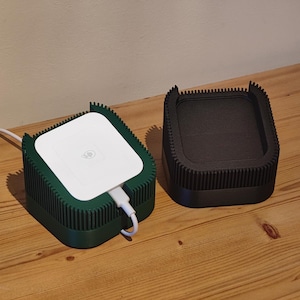 May include: Two square wireless chargers. One is dark green with a white top and a charging cable. The other is black. Both have a ribbed design around the sides and sit on a wooden surface.