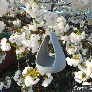 May include: A gray ceramic bird feeder shaped like a teardrop, hanging from a tree branch. The feeder has a hole in the bottom for birds to access the food. The feeder is hanging in front of a tree with white blossoms.
