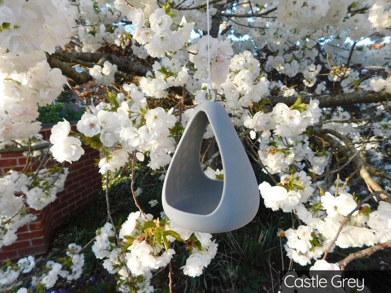 May include: A gray ceramic bird feeder shaped like a teardrop, hanging from a tree branch. The feeder has a hole in the bottom for birds to access the food. The feeder is hanging in front of a tree with white blossoms.