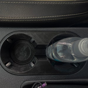 May include: A clear plastic bottle with a pump dispenser is placed in a car's cup holder. The cup holder is black and has a textured surface.