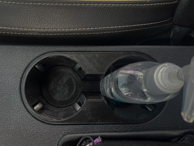May include: A clear plastic bottle with a pump dispenser is placed in a car's cup holder. The cup holder is black and has a textured surface.