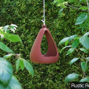May include: A rustic red triangular bird feeder hanging from a white string. The feeder is made of ceramic and has a smooth, rounded shape.