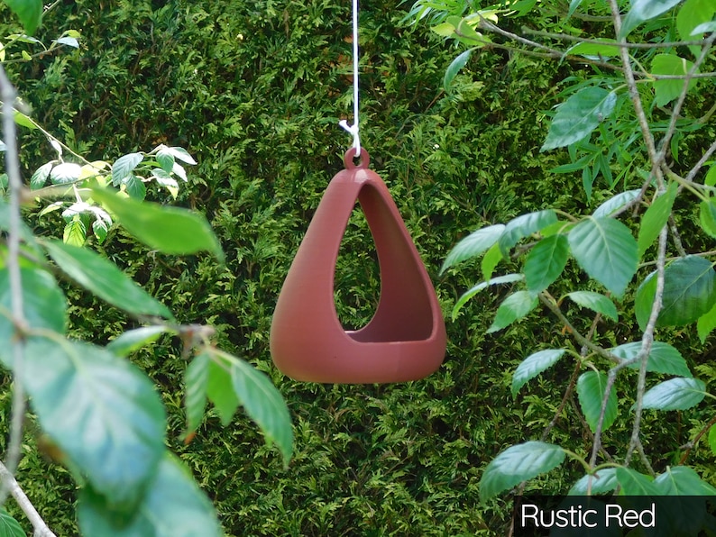 May include: A rustic red triangular bird feeder hanging from a white string. The feeder is made of ceramic and has a smooth, rounded shape.