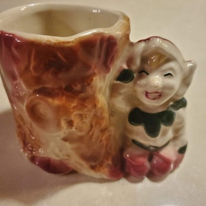 May include: A ceramic planter shaped like a tree stump with a whimsical elf figurine peeking out from behind it. The elf is wearing a green and red outfit and has a playful expression.