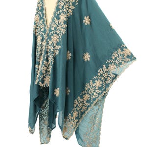 May include: Teal-colored shawl with intricate beige floral embroidery along the edges and scattered across the fabric. The shawl is draped over a mannequin, showcasing its flowing design and detailed craftsmanship. The garment appears to be made of a soft, lightweight material.