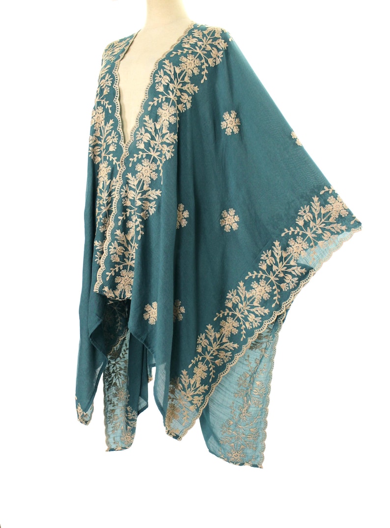 May include: Teal-colored shawl with intricate beige floral embroidery along the edges and scattered across the fabric. The shawl is draped over a mannequin, showcasing its flowing design and detailed craftsmanship. The garment appears to be made of a soft, lightweight material.