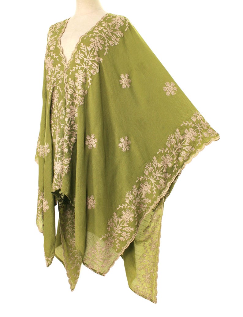 May include: Olive green embroidered cape with a V-neckline. The cape features intricate floral embroidery in a contrasting light beige color along the edges and around the neckline. The fabric appears lightweight and flowing, suitable for layering.