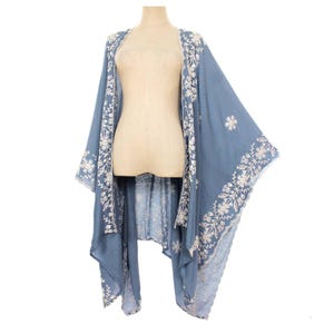 May include: A flowing, light blue kimono-style cover-up with intricate white floral embroidery along the edges and sleeves. The lightweight fabric drapes elegantly, creating a relaxed and bohemian aesthetic. A floral design is embroidered on the back.