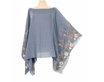 May include: A blue poncho featuring a wide boat neckline and a loose, draped silhouette. The garment is decorated with intricate floral embroidery in shades of white, yellow, and pink along the edges, creating a detailed botanical pattern.