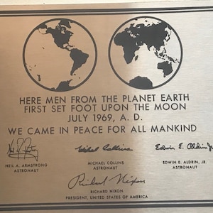Op de afbeelding: Een zilveren plaquette met een zwart-wit afbeelding van de aarde, de tekst "HERE MEN FROM THE PLANET EARTH FIRST SET FOOT UPON THE MOON JULY 1969, A. D. WE CAME IN PEACE FOR ALL MANKIND", en de handtekeningen van Neil A. Armstrong, Michael Collins, Edwin E. Aldrin Jr. en Richard Nixon.