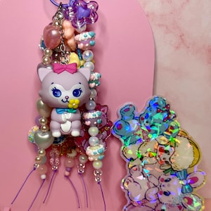 May include: A purple keychain featuring a central lavender cat figure with a pink bow and yellow flower, surrounded by various beads in shades of pink, blue, and white. Beside it is a holographic acrylic charm depicting a stack of cartoon animals.