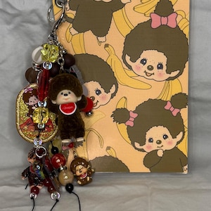 May include: A keychain with a brown plush monkey charm, various beads, and a notebook featuring cartoon monkeys and bananas. The notebook has a clear plastic cover and a silver metal ring. The background is a light yellow.
