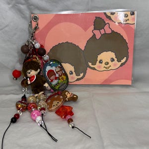 May include: A keychain with multiple charms and beads, including a brown plush figure, a small oval charm with a cartoon illustration, and a pink and red beaded strand. A pink card with a cartoon character illustration is in the background.