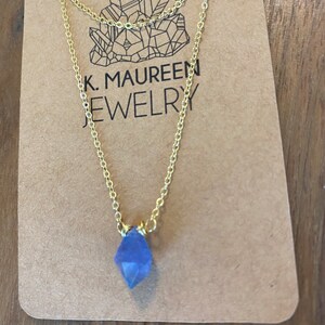 May include: A delicate gold chain necklace with a small, faceted, light blue crystal pendant. The necklace is displayed on a brown card with the text "K. MAUREEN JEWELRY".