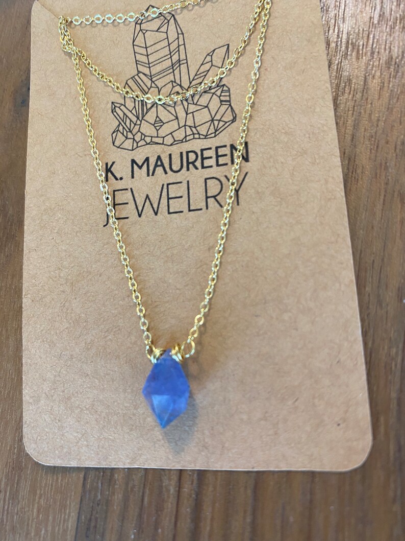 May include: A delicate gold chain necklace with a small, faceted, light blue crystal pendant. The necklace is displayed on a brown card with the text "K. MAUREEN JEWELRY".