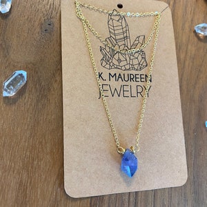May include: A gold chain necklace with a blue crystal pendant. The necklace is layered and has a delicate design. The pendant is a faceted crystal with a pointed shape. The necklace is displayed on a brown card with the text "K. Maureen Jewelry".